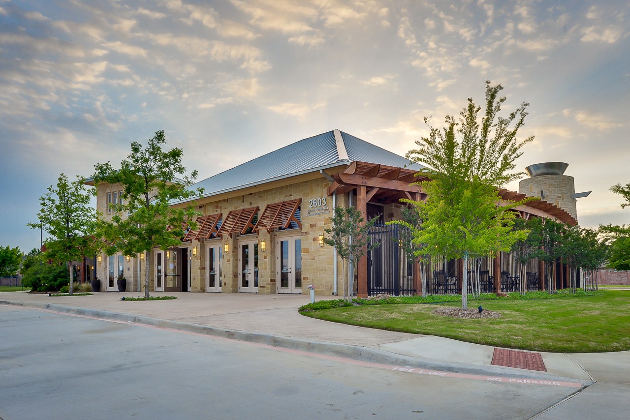 A single-story limestone community building with a standing-seam metal roof, a row of glass double doors under slatted cedar awnings, a covered pergola entry at one end and a round stone tower at the other, with young trees and a lawn along the fire lane out front.