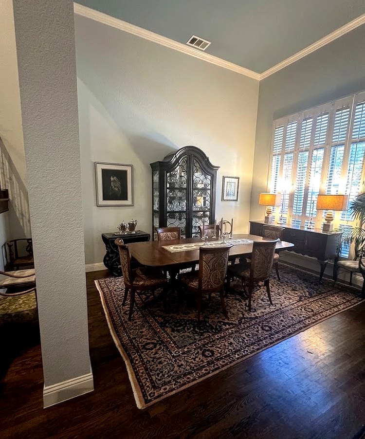 The former formal living room in use as the dining room. A table with six upholstered chairs stands on a large patterned rug with room to walk all the way around it, below a high crown-moulded ceiling, with a glass-fronted china cabinet and framed pictures on the far wall and a lamp-topped sideboard beside tall plantation-shuttered windows.