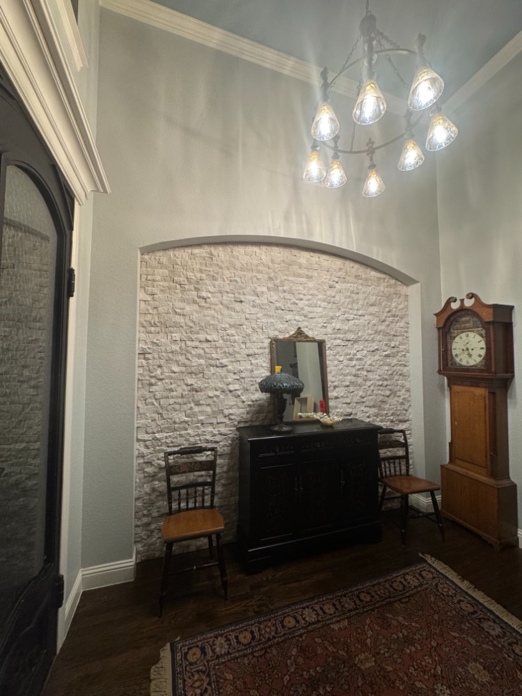 The same arch after it was closed in, now a recessed panel of pale stacked stone facing the foyer. A black console table with a lamp and a gilt mirror stands against it, flanked by two wooden chairs, with a tall longcase clock to the right.