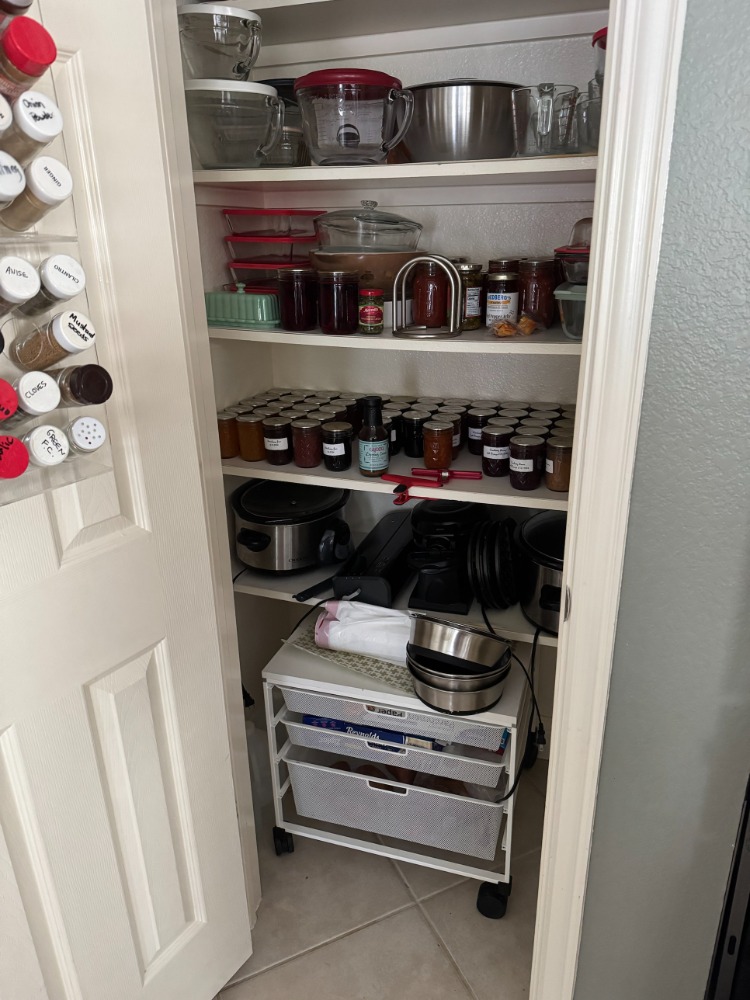 The original reach-in pantry closet, about one door wide, with five fixed shelves packed to the edges with mixing bowls, home-canned jars, slow cookers and bakeware, plus a spice rack mounted on the back of the door and a rolling wire drawer unit standing on the floor.