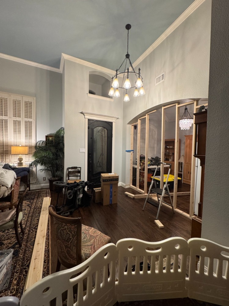 The arched dining room opening part way through construction, framed in with new vertical lumber studs. A step ladder, lumber offcuts and moving boxes sit on the floor of the entry, with the front door and the chandelier visible alongside.