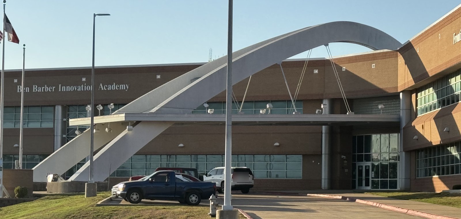 A long two-story brick school building lettered Ben Barber Innovation Academy, fronted by a sweeping white steel arch that rises over the entrance canopy, with flagpoles at the far left and vehicles parked along the drive.