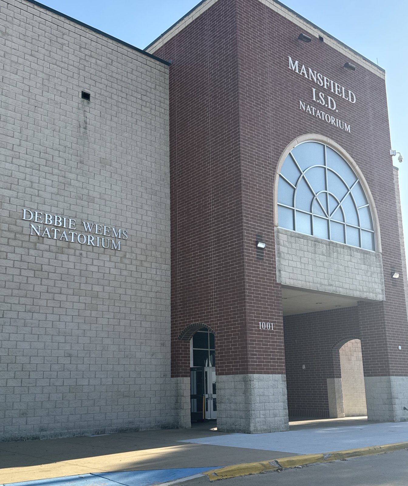 The corner of a dark brick and grey block building with a large fan-shaped arched window above the entry, lettered Mansfield I.S.D. Natatorium on the brick face and Debbie Weems Natatorium on the block wall beside the doors.