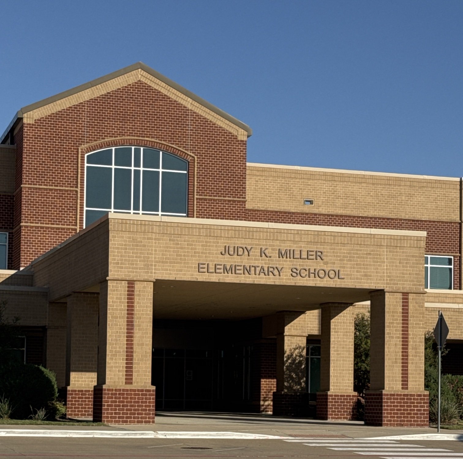 The front of a brick elementary school, with a tan brick drop-off canopy carrying the name Judy K. Miller Elementary School in raised letters, a tall arched window in the red brick gable above, and a crosswalk striped across the drive in the foreground.