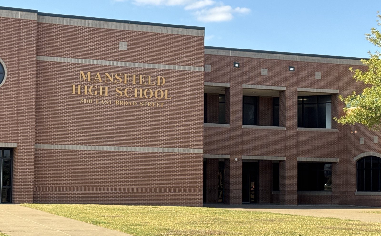 A two-story red brick school building with Mansfield High School, 3001 East Broad Street, spelled out in gold letters across a wide brick panel, with tall dark windows and a columned entrance bay to the right under a clear blue sky.