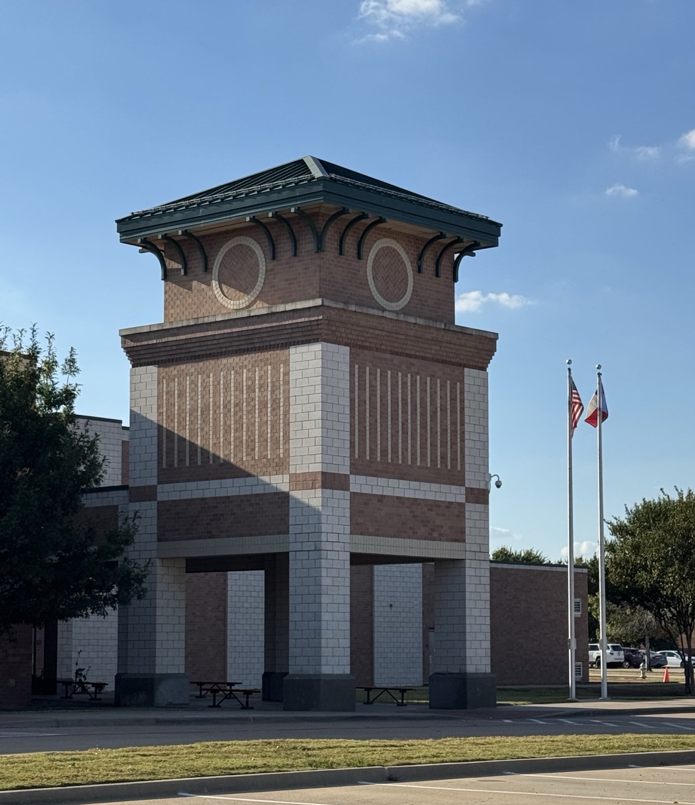 A tall square entry tower of red brick and cream block, topped with a green hipped metal roof and circular brick medallions, standing open at ground level over picnic tables, with United States and Texas flags on poles to the right.