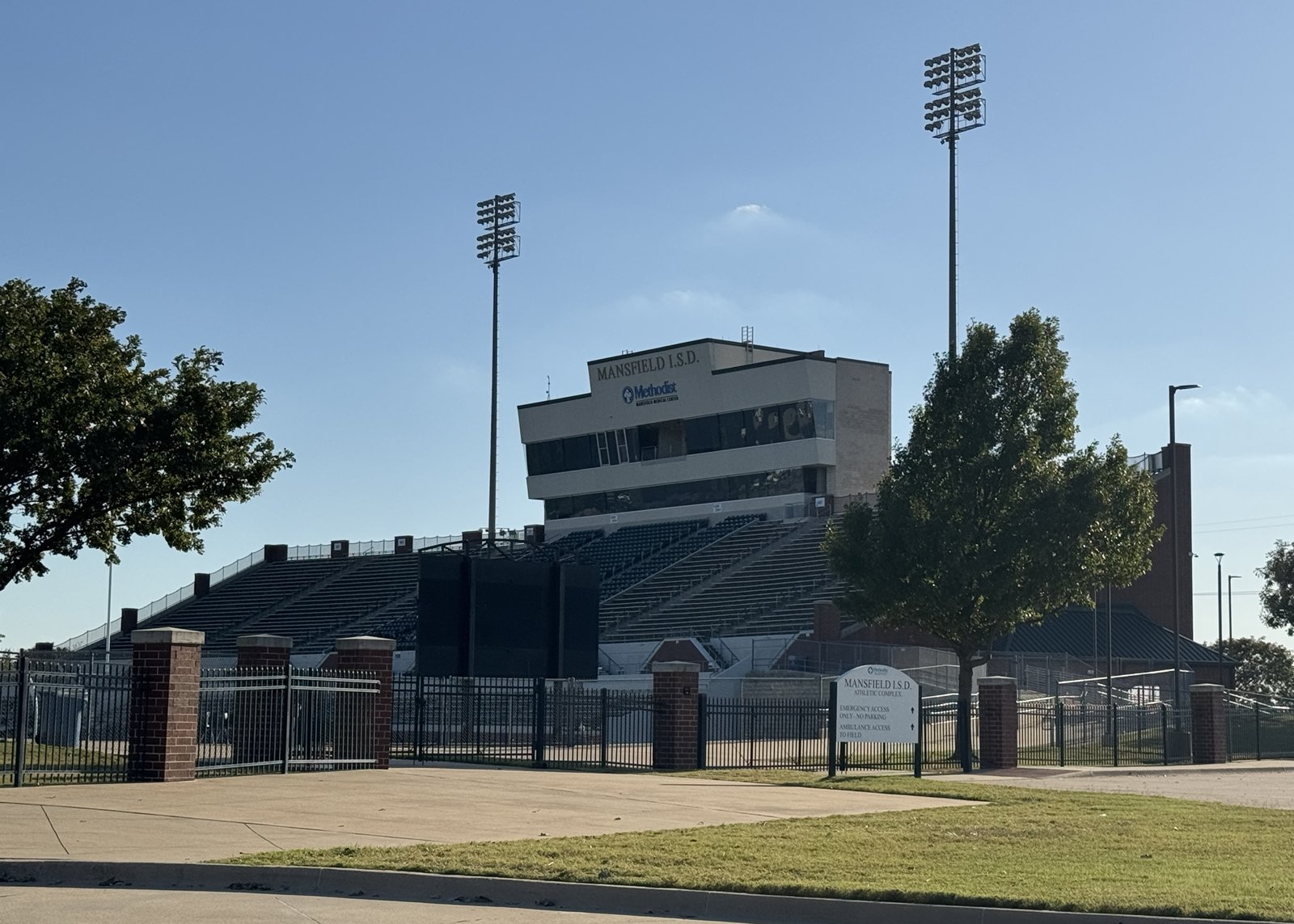 A high school football stadium seen from beyond its iron fence, with banked metal bleachers, a multi-level press box lettered Mansfield I.S.D. and two tall floodlight towers; a sign at the gate reads Mansfield I.S.D. Athletic Complex.