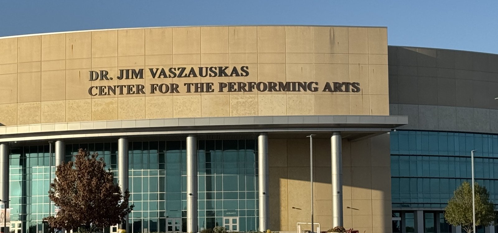 A gently curved limestone-clad building front lettered Dr. Jim Vaszauskas Center for the Performing Arts above a run of tall windows and slender metal columns, with a small red-leaved tree beside the entrance.