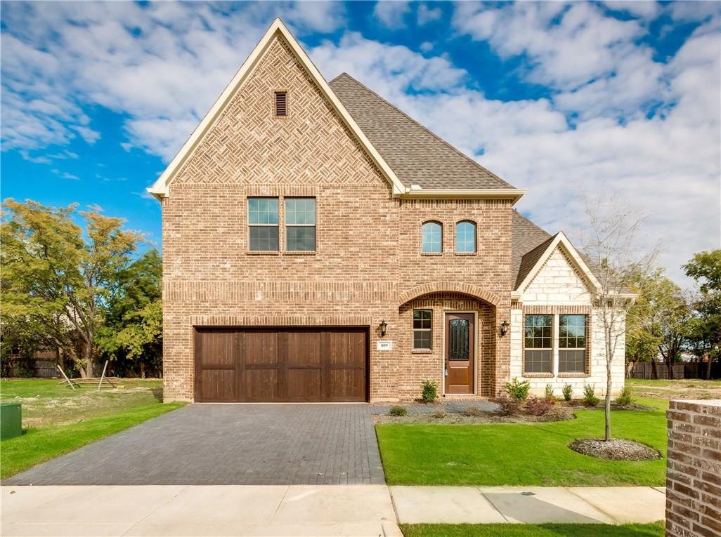 A two-story brick home with a wooden garage door and a paver driveway, with a low brick wall and fencing marking the edges of the front lot.